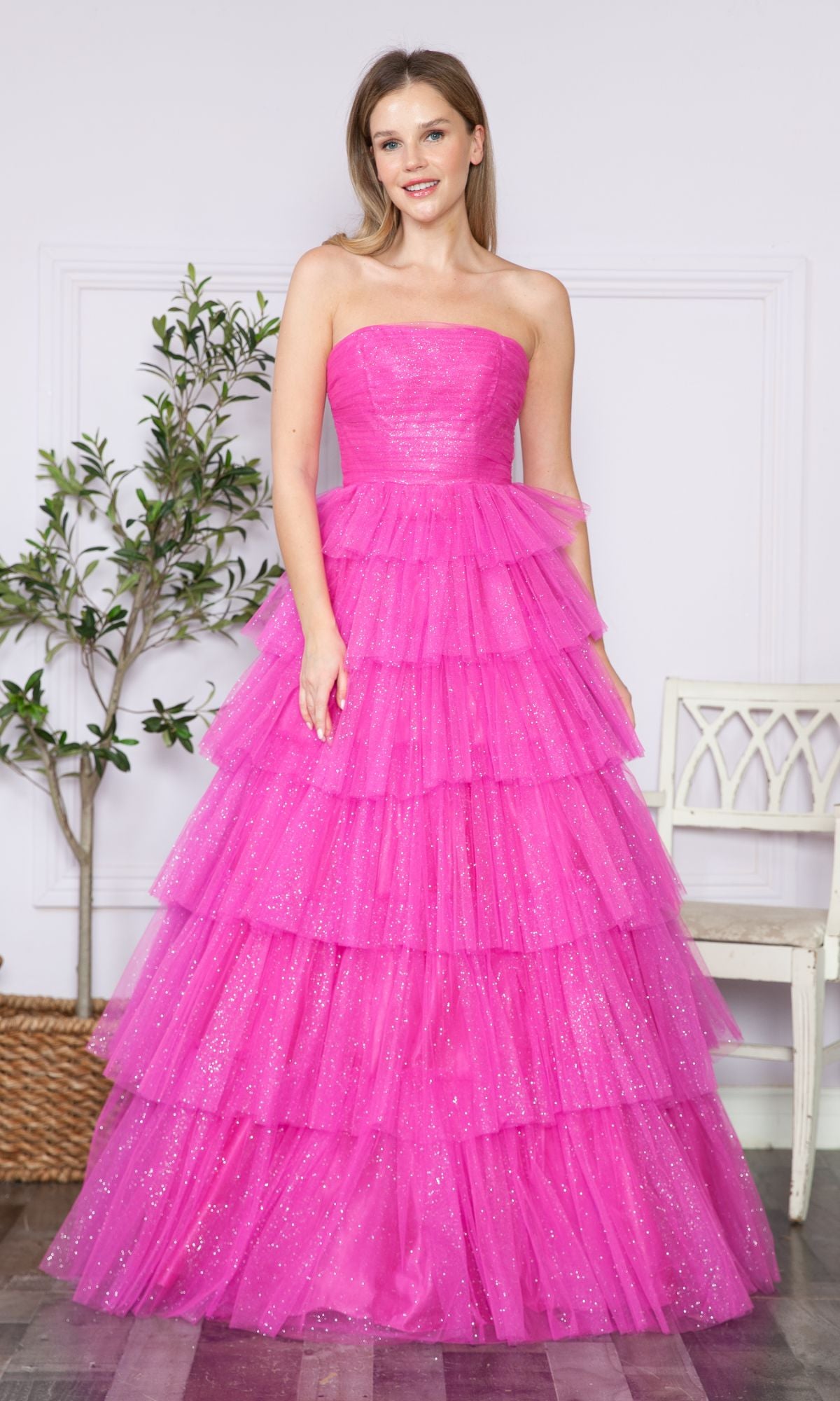 A woman stands indoors wearing the Strapless Long Tiered Glitter Prom Ball Gown 9386 in bright pink tulle with multiple ruffled layers, set against a simple background featuring a plant and a white chair.